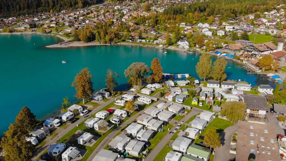 Vue aérienne d’un camping avec mobil-homes et caravanes au bord d’un lac turquoise, entouré de maisons et de montagnes boisées