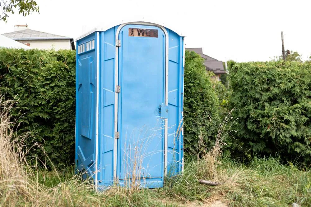 Cabine de toilettes sèches mobiles bleue installée en extérieur, au bord d’une haie et d’un terrain enherbé