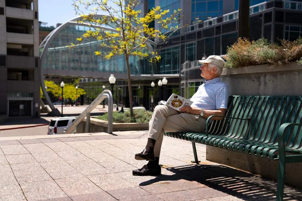 Homme senior assis sur un banc public en ville, lisant un magazine par une journée ensoleillée.